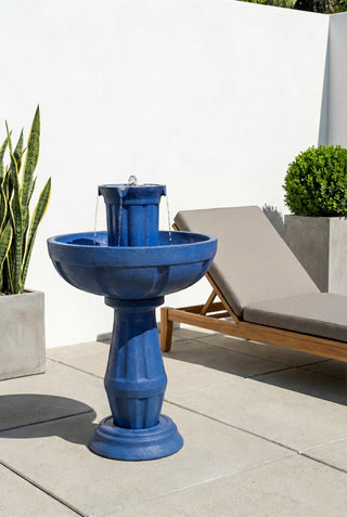 Blue outdoor fountain on a patio with a lounge chair and plants in the background