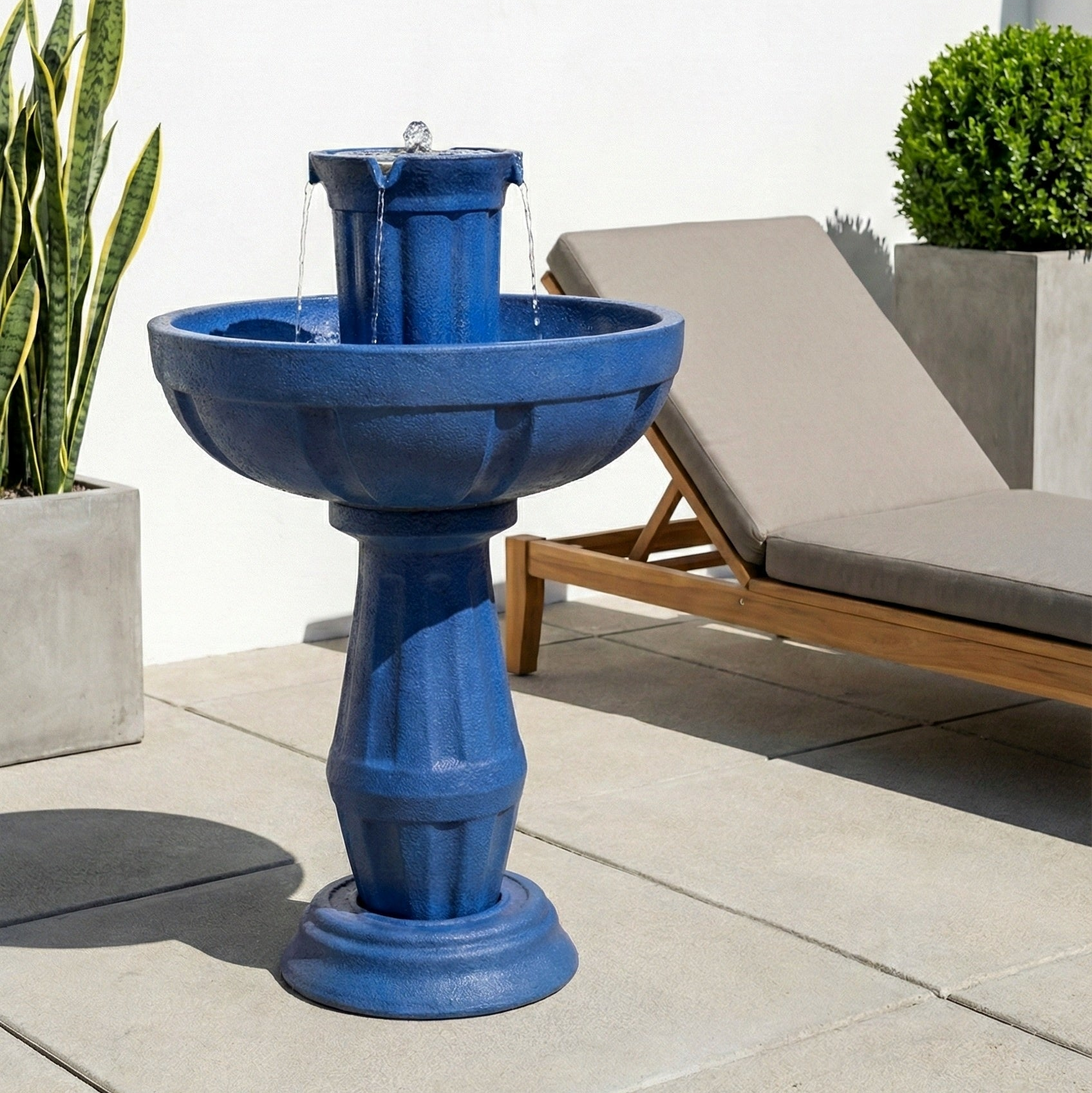Blue outdoor fountain on a patio with plants and a lounge chair in the background