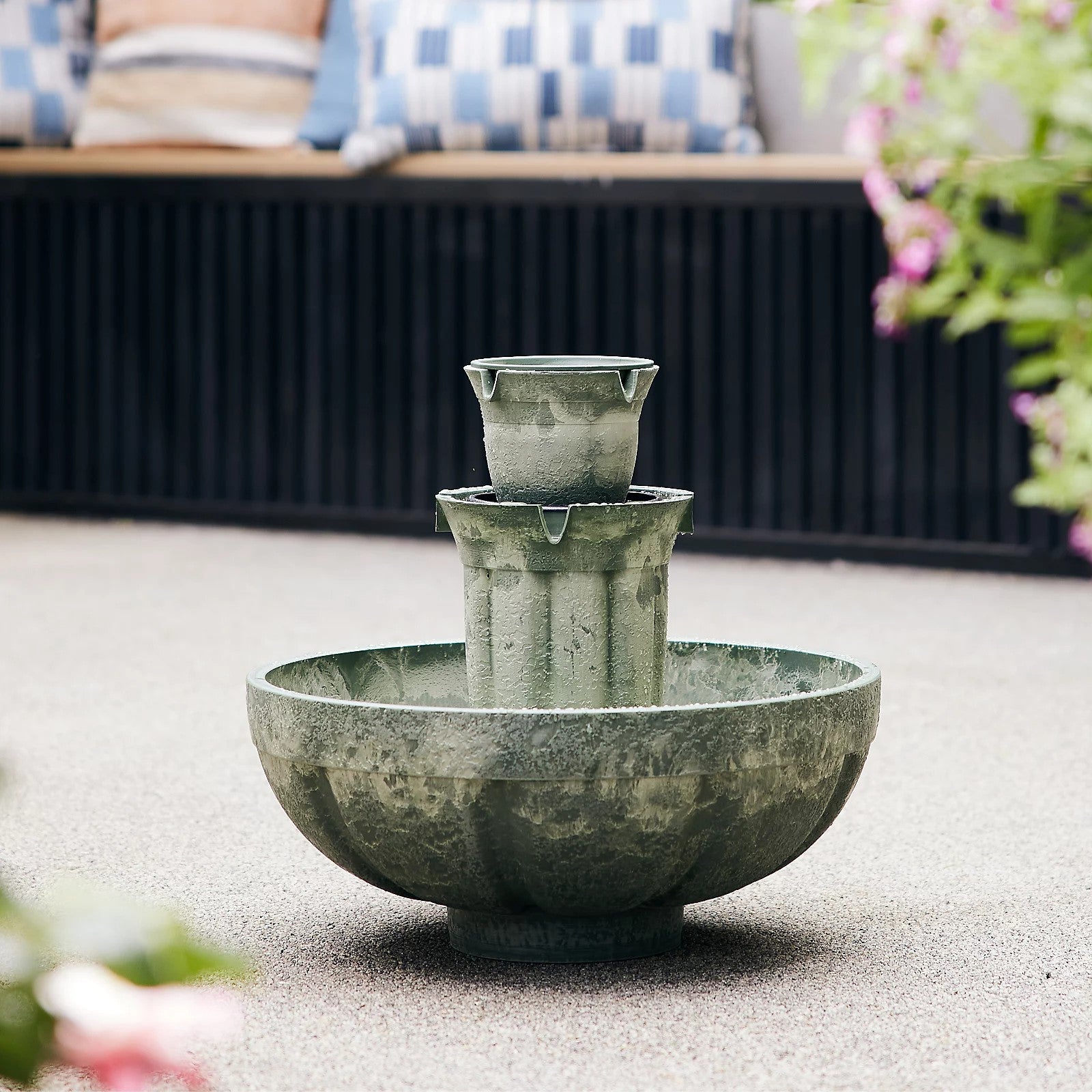 Outdoor fountain with a rustic design on a concrete surface, with a blurred background of outdoor furniture and plants.