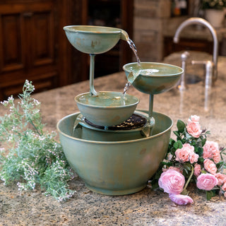 Small green ceramic water fountain on a kitchen counter with flowers and plants.