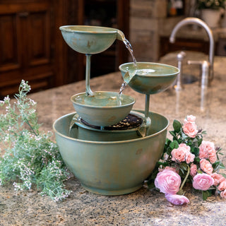 Small green ceramic water fountain on a kitchen counter with flowers and plants.