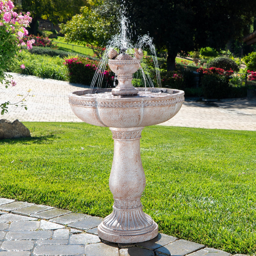 Decorative outdoor fountain in a garden setting with greenery and a building in the background.