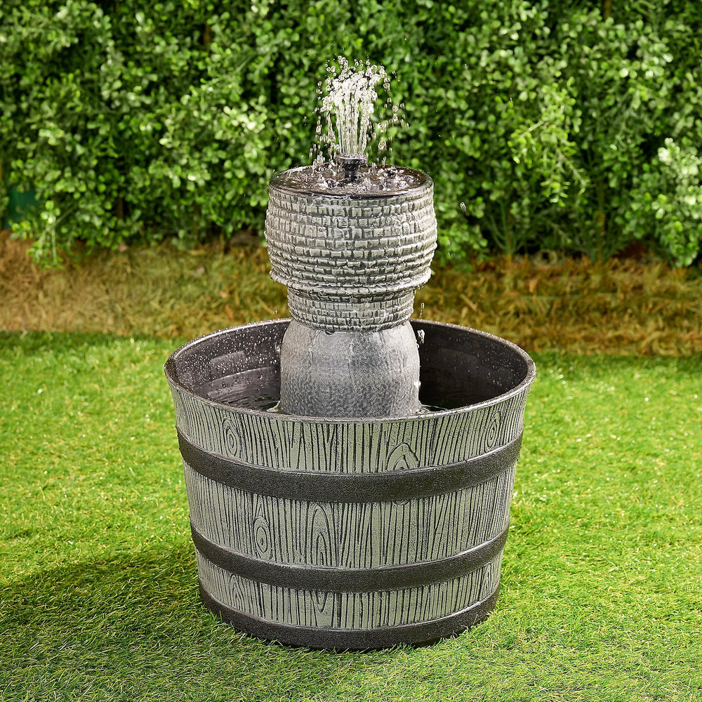 A rustic whiskey barrel-shaped fountain with a gray finish, placed on green grass against a backdrop of green shrubbery.