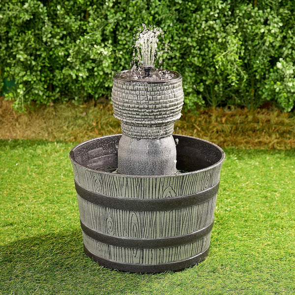 A rustic whiskey barrel-shaped fountain with a gray finish, placed on green grass against a backdrop of green shrubbery.