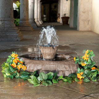 Fountain with water feature surrounded by greenery and flowers in a stone courtyard.